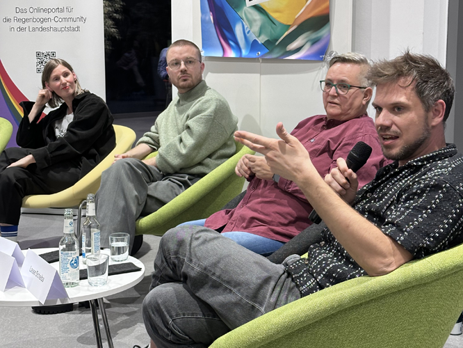 Vier Personen sitzen auf einer Bühne in bequemen Sesseln und diskutieren bei einer Veranstaltung; eine Person spricht gerade in ein Mikrofon.