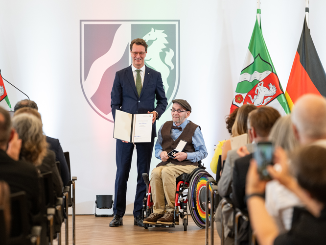 Ein Mann in Anzug überreicht einer im Rollstuhl sitzenden Person eine Urkunde auf einer Bühne. Im Hintergrund sind die Flaggen von NRW und Deutschland sowie ein Wappen mit einem springenden Pferd zu sehen. Im Vordergrund sitzt ein Publikum.