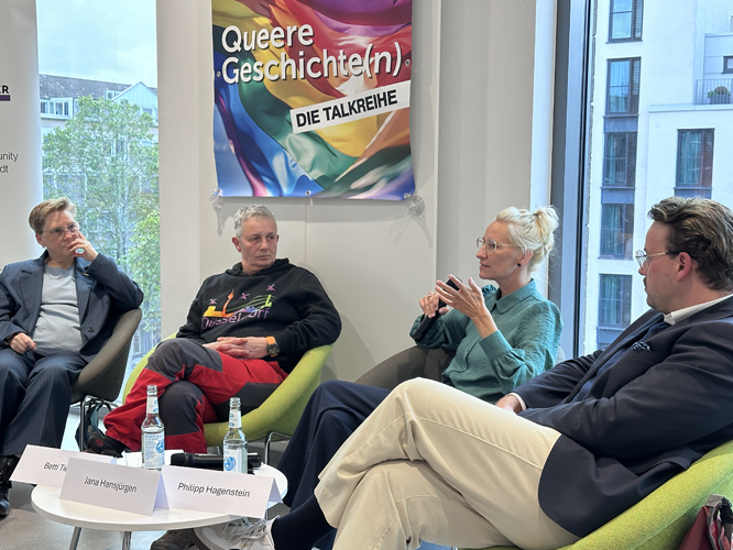 Vier Personen sitzen auf grünen Sesseln bei einer Podiumsdiskussion vor einem Banner mit der Aufschrift Queere Geschichte(n) – DIE TALKREIHE und einer Regenbogenflagge. Im Hintergrund sind große Fenster mit Blick auf Bäume und Stadtgebäude zu sehen.