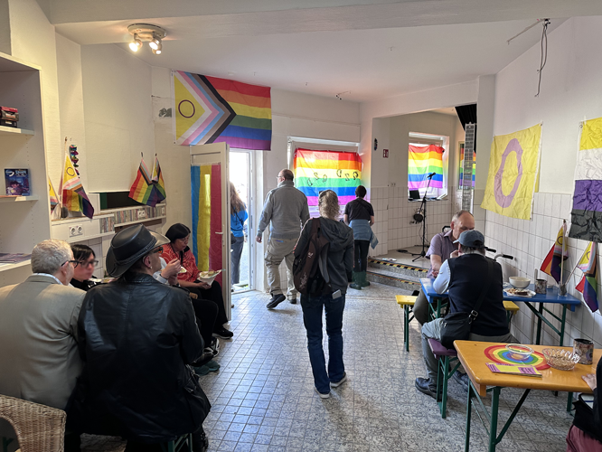 Innenraum des Queeres Zentrum Düsseldorf mit mehreren Personen, die sich unterhalten oder an Tischen sitzen. An den Wänden hängen Regenbogenflaggen und bunte Banner.