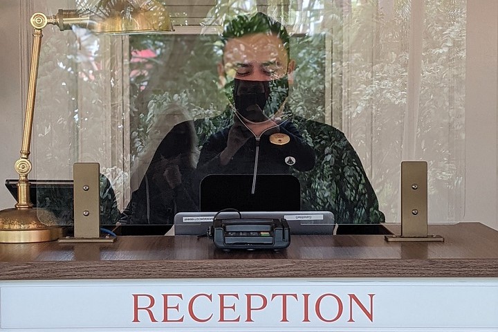 Ein Mann mit Maske steht hinter der Hotelrezeption mit Plexiglas als Synonym für die Initiative #perspektivejetzt