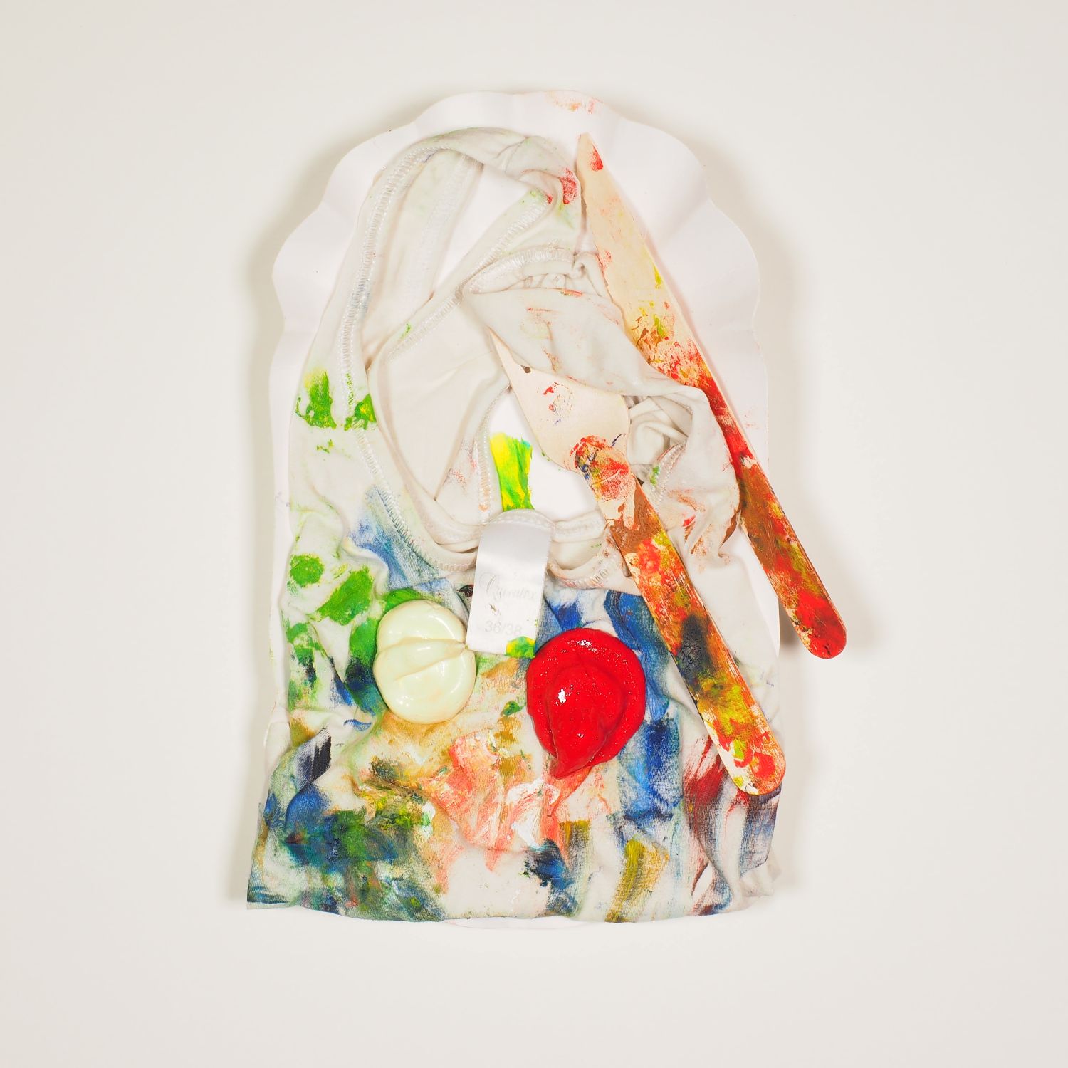 Assemblage von einem Pommesschälchen, einem Unterhemd voller Farbflecken, je einem Klecks Ketchup und Majo und Essbesteck