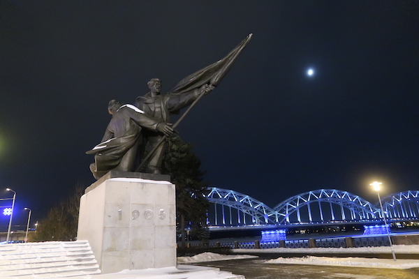 Revolutionsdenkmal 1905 in Riga bei Nacht mit angestrahlter Brücke im Hintergrund