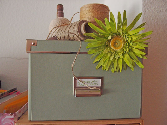 Schachtel mit grüner Blume, Mentallbeschlägen und Garnrollen auf einem Schrank neben Ordnern