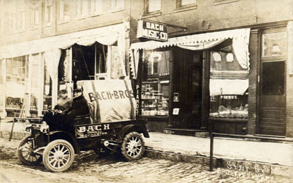 Historische Bach-Genealogie: ein alter Kleinst-LKW auf einem schwarzweißem Foto sthet vor einem Ladengeschäft in den USA. Auf dem vorderen unüberdachten Sitz sitzen 2 Personen. Am Ausleger sieht man den Schriftzug Bach, ebenso auf dem Auto.