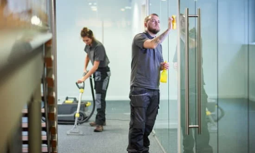 Büroreinigung für die Region Karlsruhe - Mann reinigt die Glasstüren des Konferenzraum, und eine Frau im Hintergrund saugt den Boden des Raumes