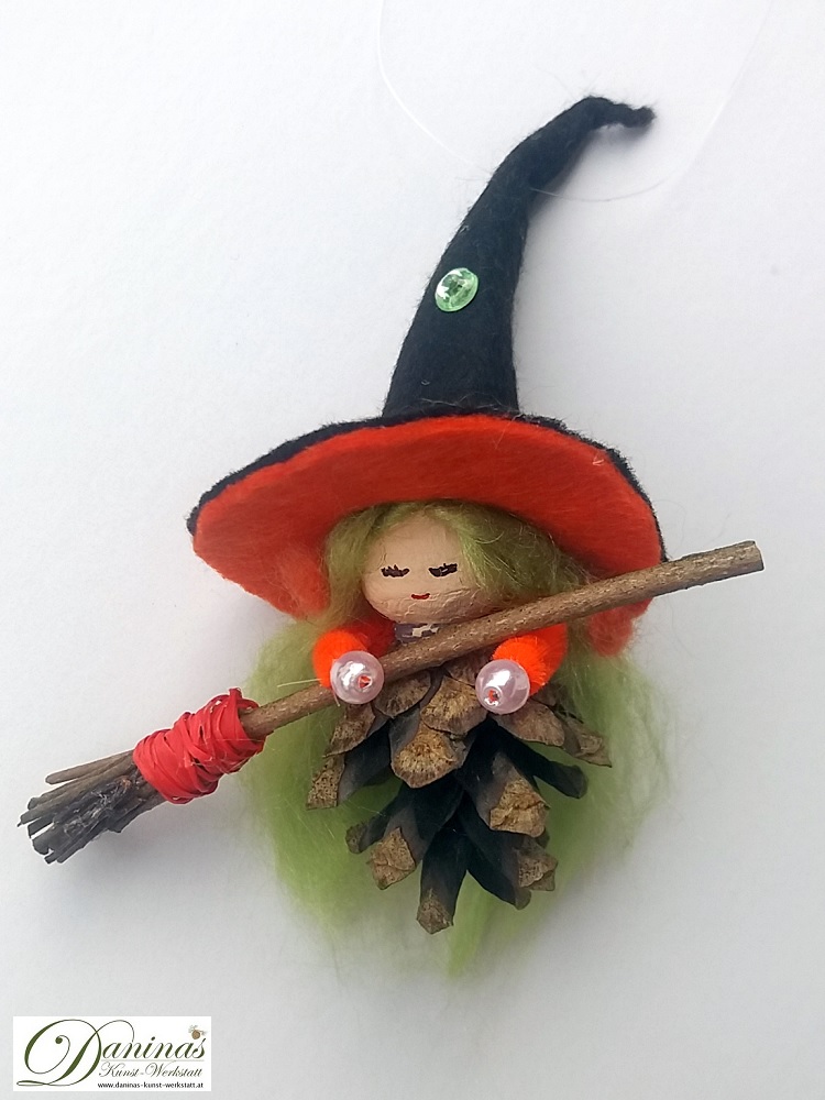 Hexe Hella. Handgefertigte Märchenfigur aus Kiefernzapfen, mit grünen langen Haaren aus Wolle, einem schwarz-orangen Hexenhut aus Filz mit einem magischen grünen Stein und einem Hexenbesen aus Zweigen