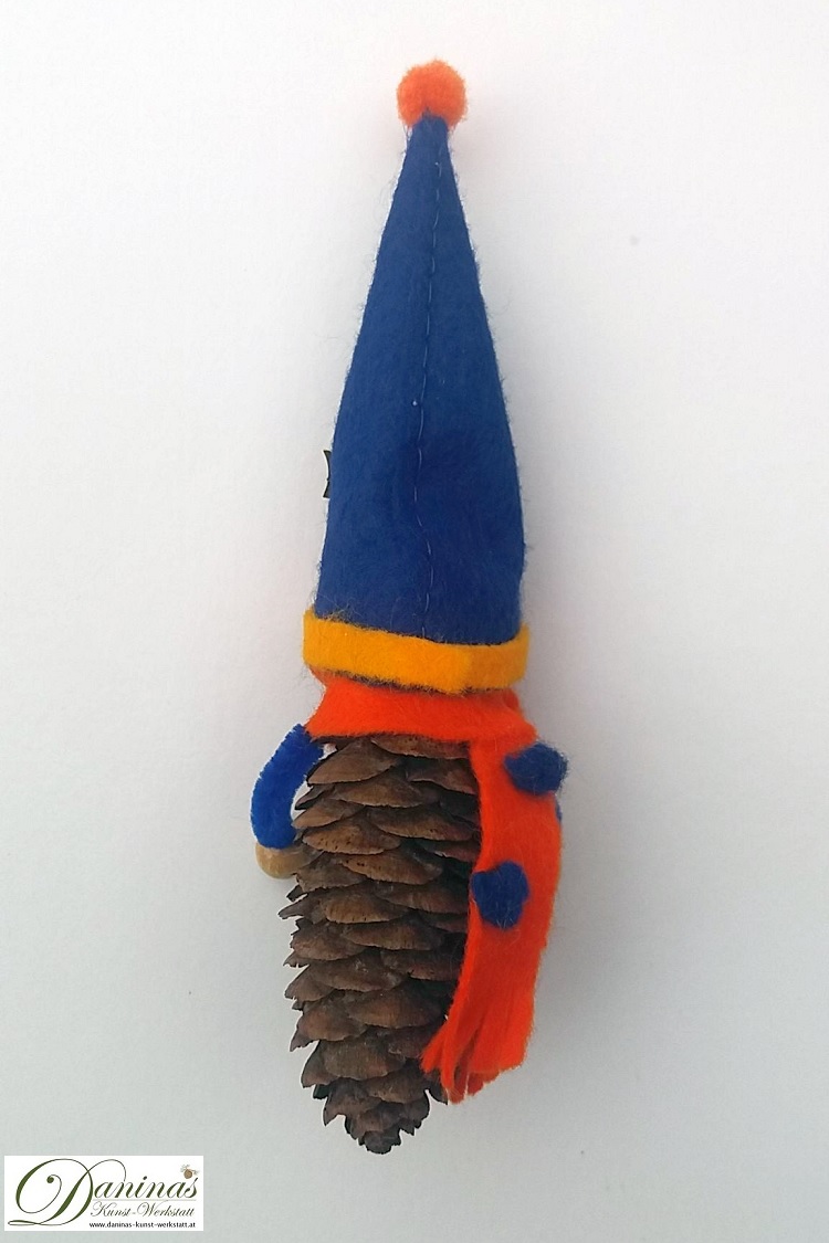 Geburtstagswichtel Benno, Rückseite. Handgefertigte Märchenfigur aus Fichtenzapfen, mit orangem Schal und blauer Zipfelmütze aus Filz mit blau-orangen Bommeln