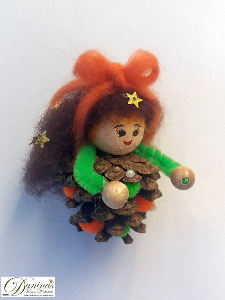 Kleine Hexe Stella. Handgefertigte Märchenfigur aus Kiefernzapfen, mit braunen langen Haaren aus Wolle mit Sternen und einem orangen Haarband, grüne und orange Bommeln im Zapfen