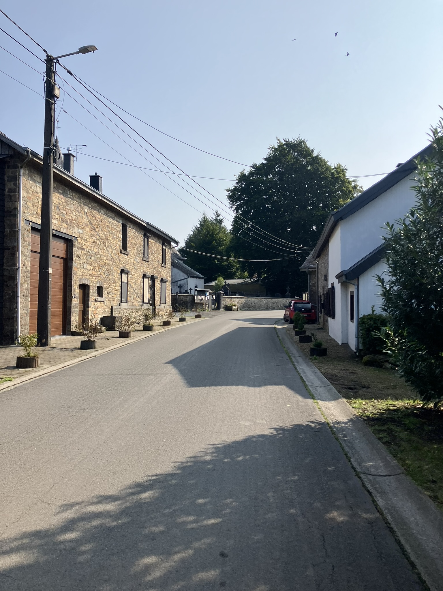 Bikepacking Belgien - Belgische Dorfstraße mit Natursteinhäusern und weißem Wohnhaus, Stromleitungen über der Straße, sonniger Tag.