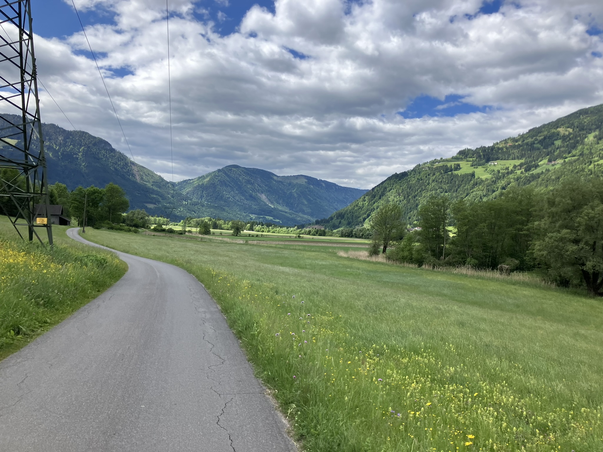 Bikepacking-Tour durch Kärnten – Drautal bei Kleblach, Österreich – Asphaltstraße zwischen Wiesen und bewaldeten Bergen unter dramatischem Wolkenhimmel.