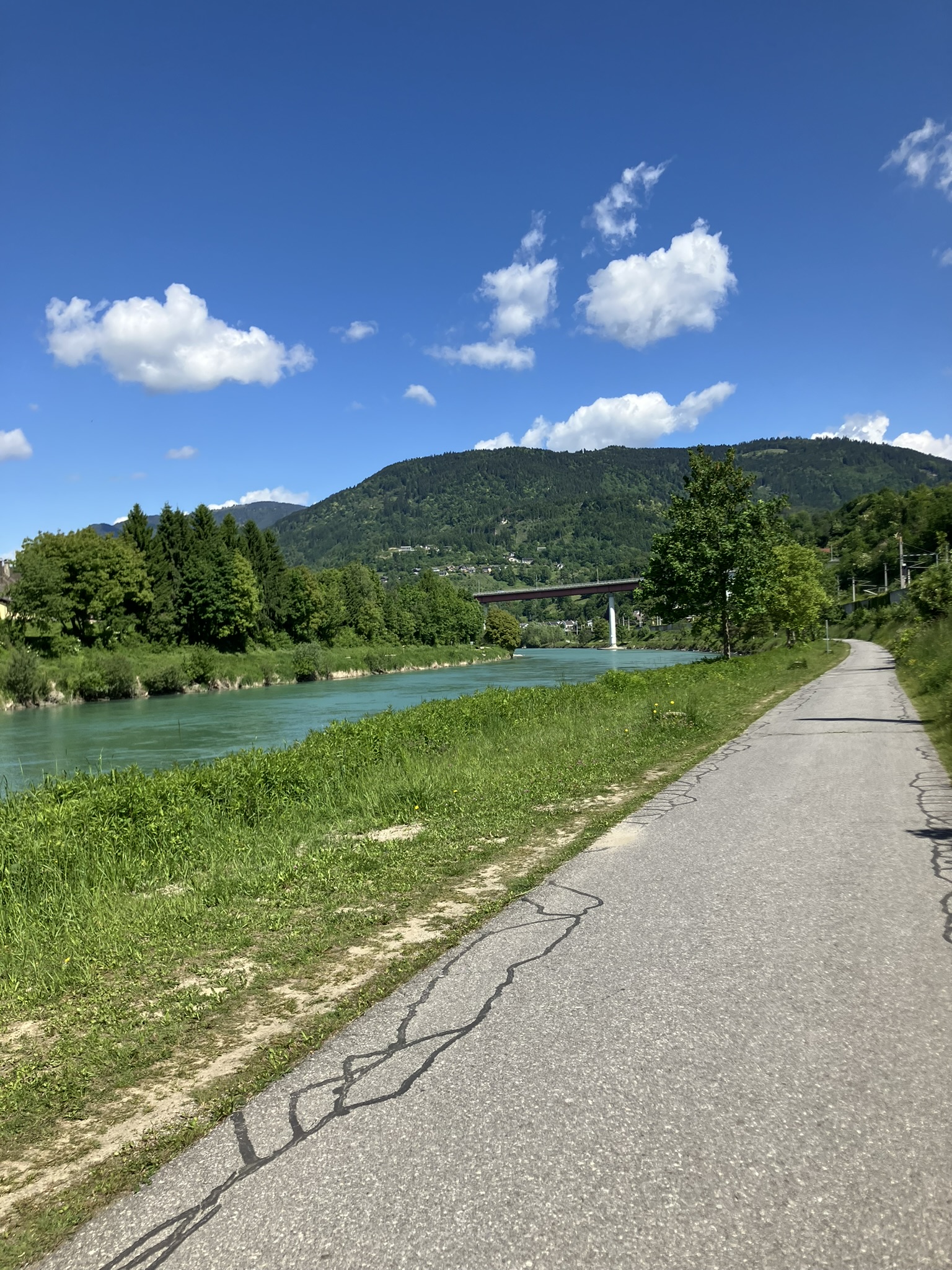 Bikepacking auf Radweg entlang der Drau in Kärnten mit Fluss, Brücke und bewaldeten Bergen im Hintergrund.