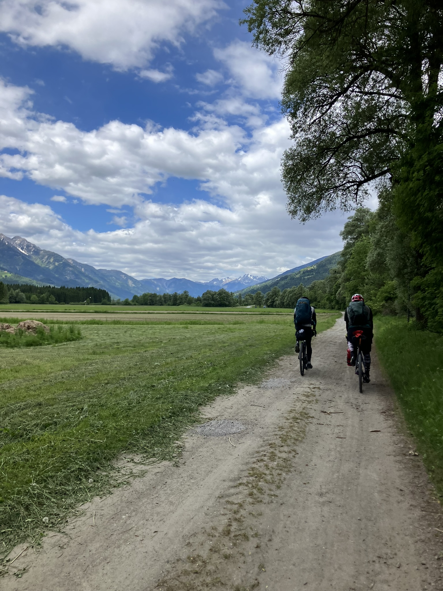 Zwei Bikepacker auf Schotterweg im Drautal in Kärnten mit Blick auf Alpen und weite Wiesenlandschaft.
