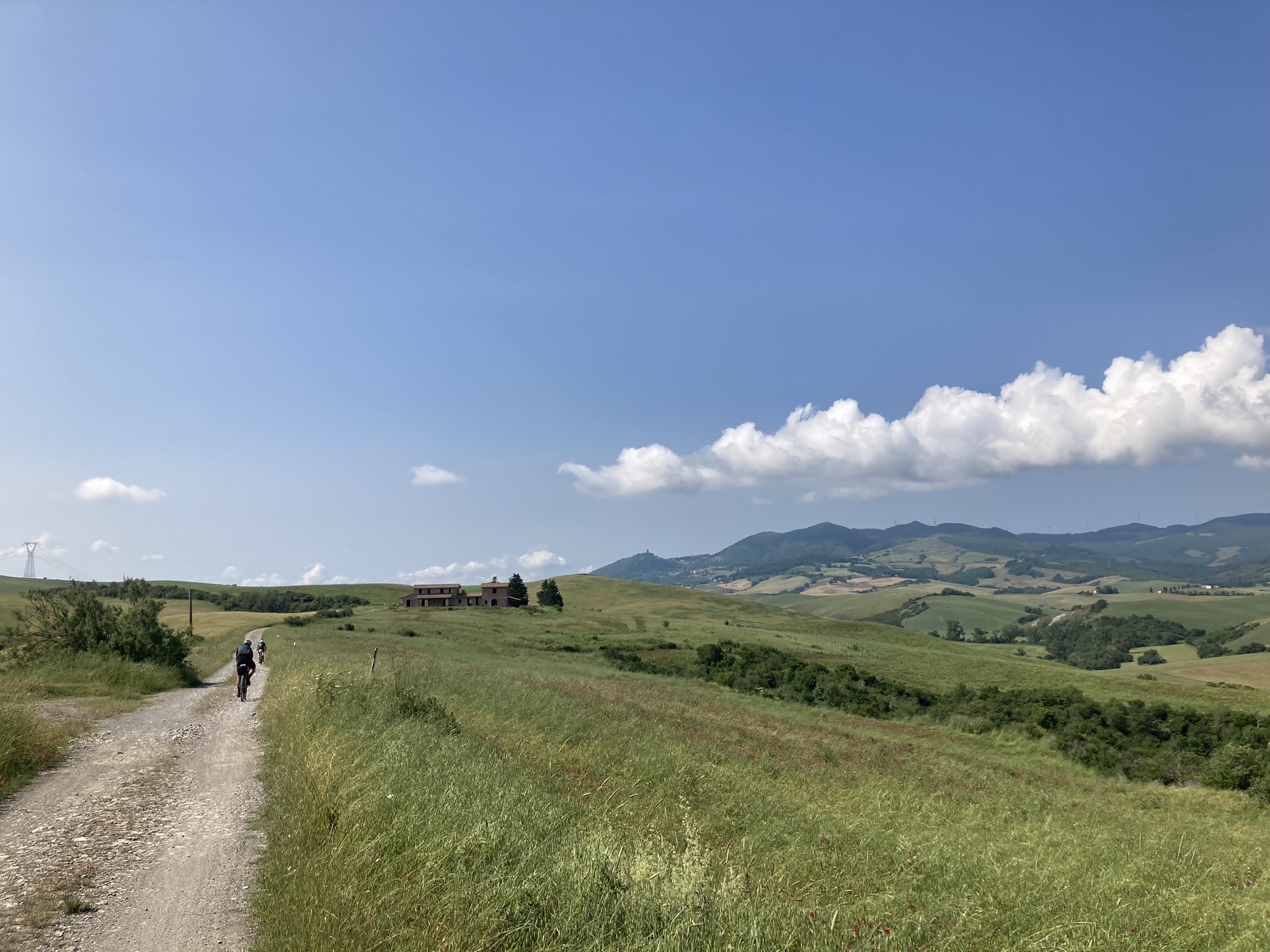 Bikepacking Italien - von Livorno nach Rimini: Radfahrer auf Schotterpiste durch offene Hügellandschaft, Felder und Berge im Hintergrund, leicht bewölkter Himmel.