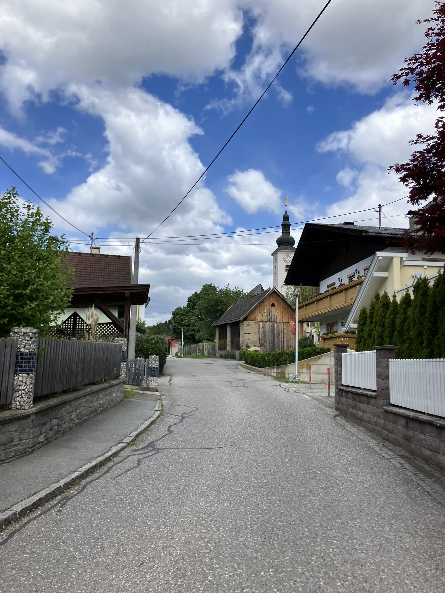 Bikepacking auf der Tour durch Kärnten, Österreich – ruhige Dorfstraße mit Holzhäusern und Kirchturm im Hintergrund, Asphalt, wechselhafte Bewölkung.