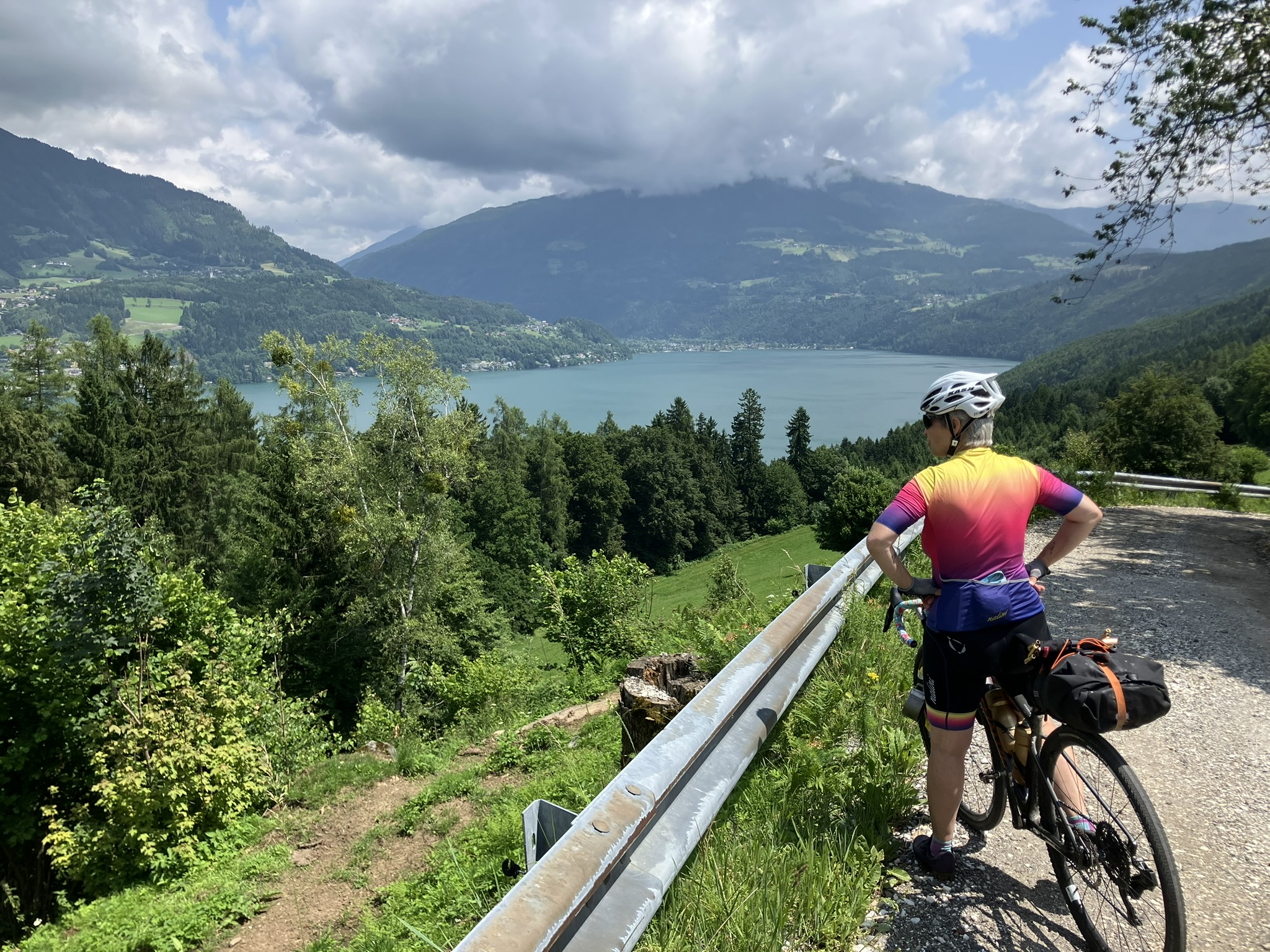 Bikepacking auf der Tour durch Kärnten, Österreich – Fahrerin mit Gravelbike am Aussichtspunkt über dem Millstätter See, Bergpanorama, Schotterstraße, dramatische Wolken.