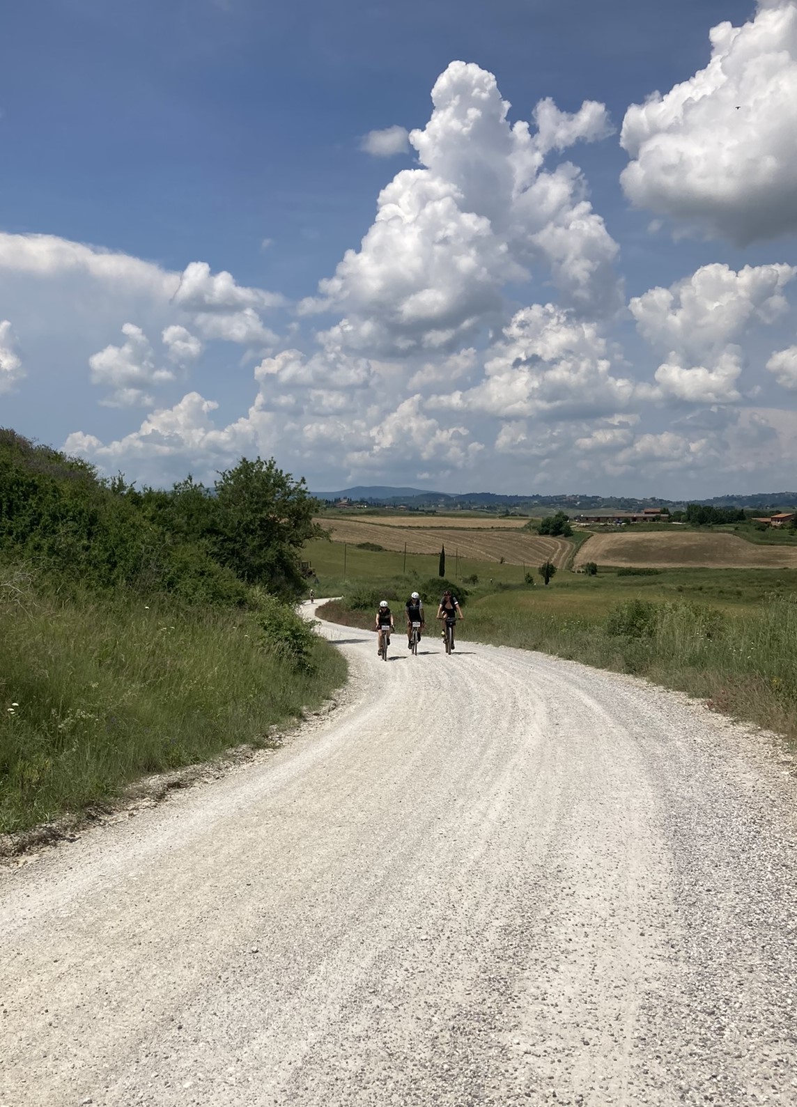 Bikepacking Italien - Toskana, helle Schotterstraße durch offene Hügellandschaft mit drei Radfahrern, aufgenommen während einer Gravel-Bikepacking-Etappe in Italien.