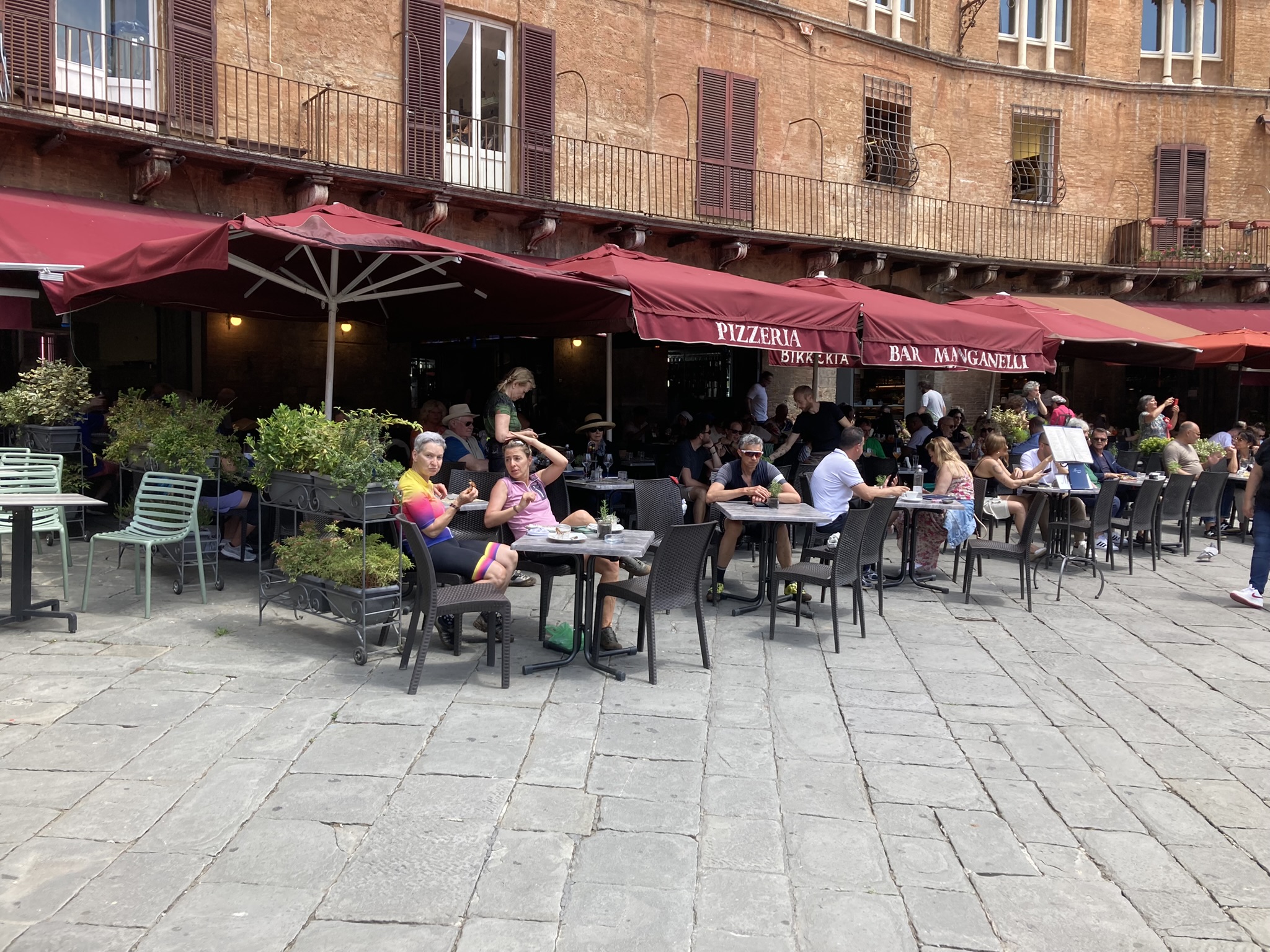 Bikepacking Italien - Stopp in Siena, Toskana: Straßencafé mit roten Sonnenschirmen an der Piazza del Campo, Radreisende genießen Espresso vor mittelalterlicher Kulisse.