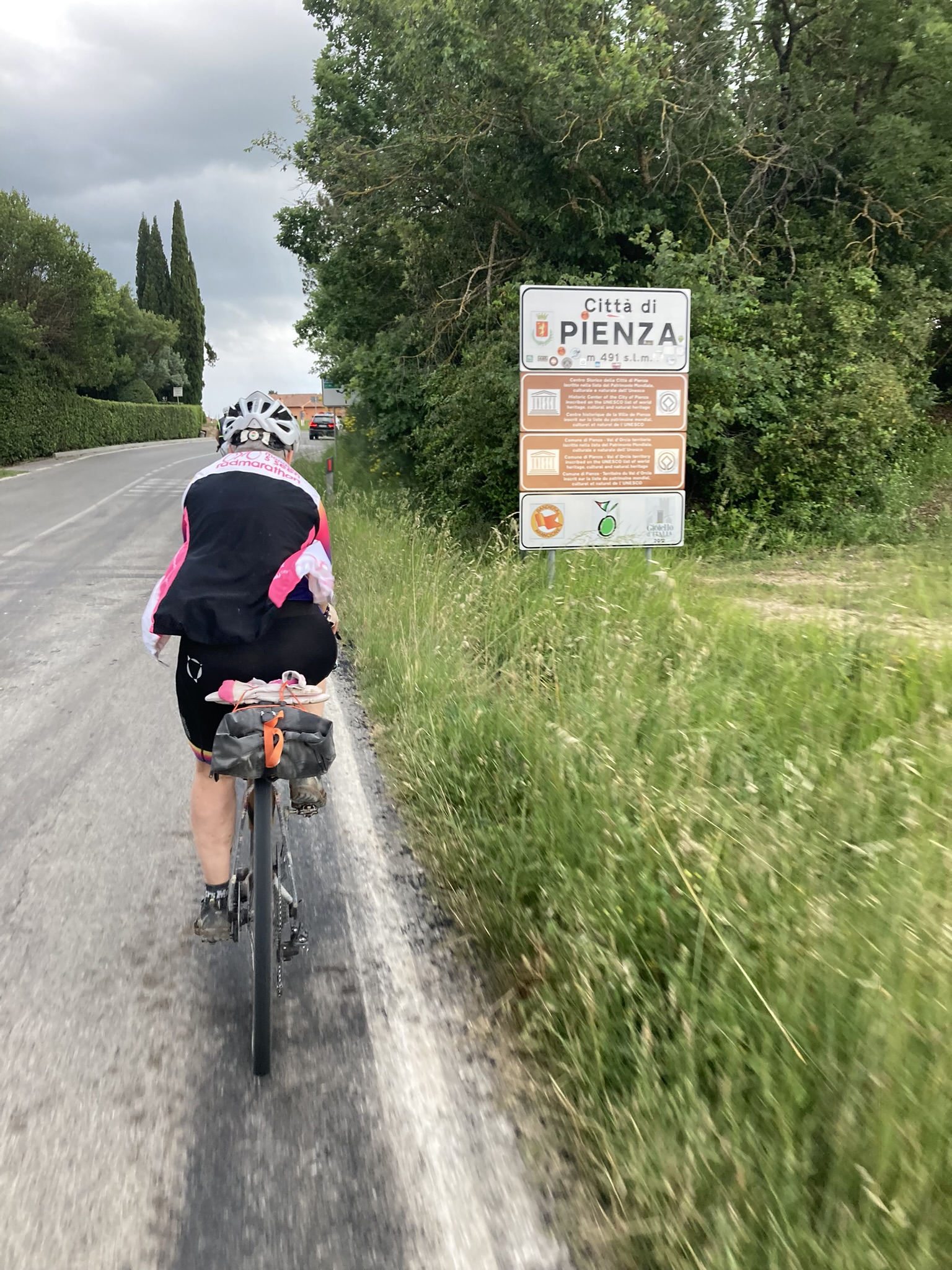 Bikepacking Italien - von Livorno nach Rimini: Radfahrerin neben Ortsschild von Pienza, Landstraße mit Zypressen und Grünflächen, bewölkter Himmel.