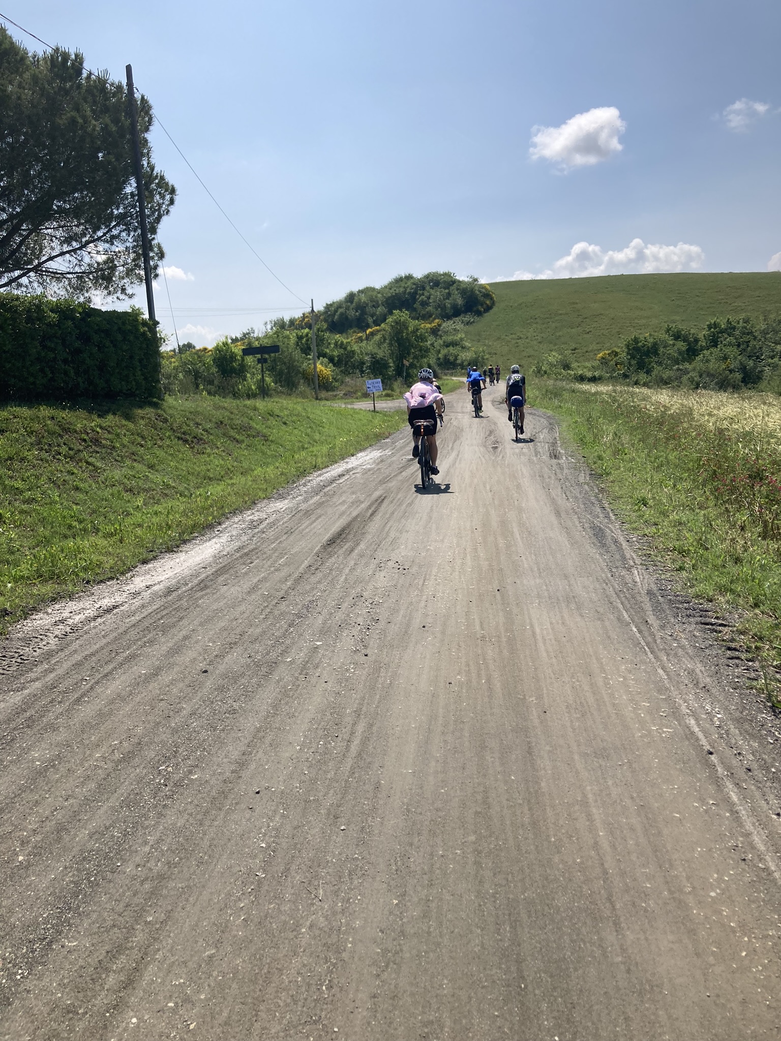 Bikepacking Italien - von Livorno nach Rimini – Toskana – Gruppe von Radfahrern auf ansteigender Schotterstraße – offene Hügellandschaft unter blauem Himmel