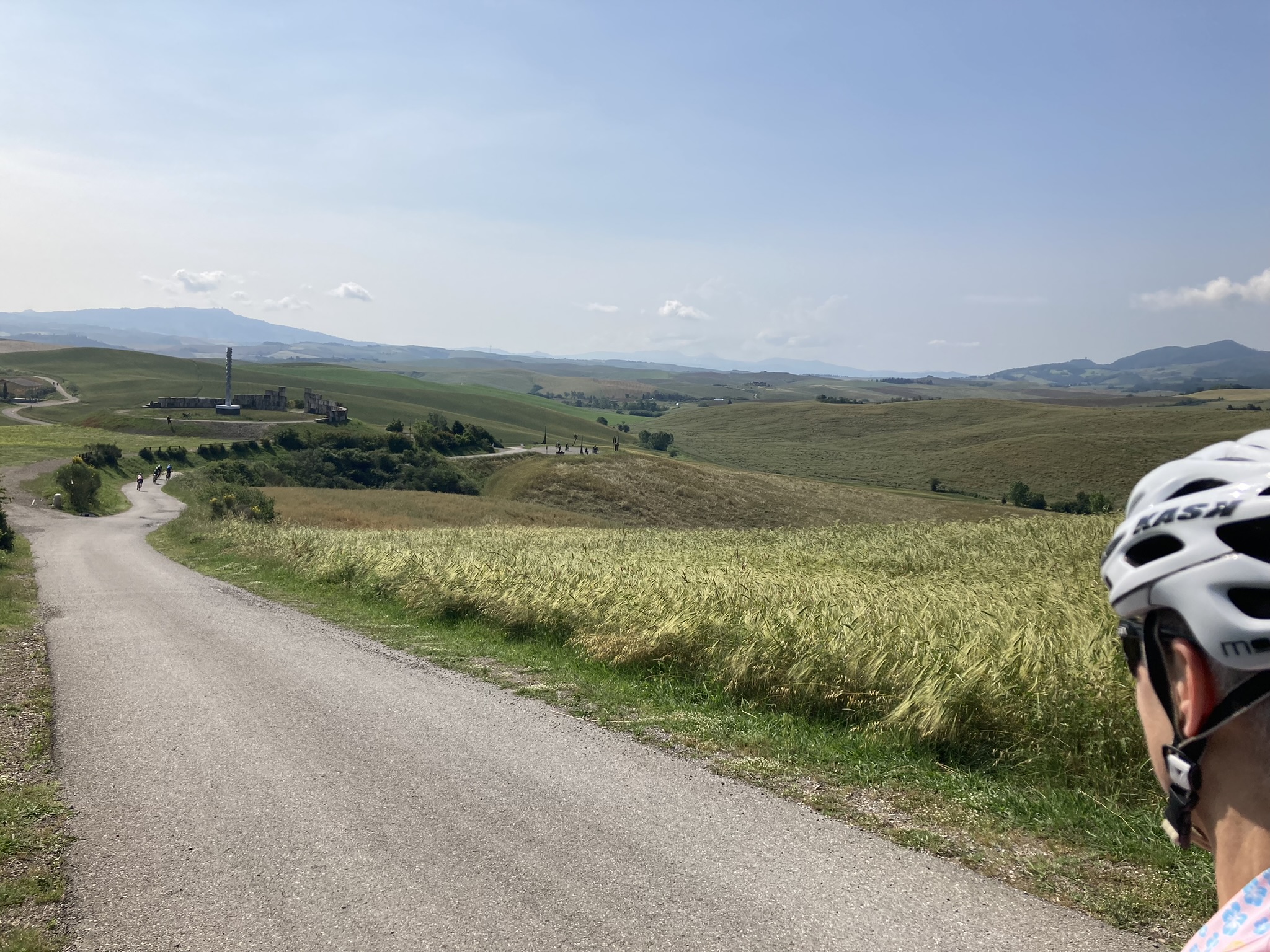 Bikepacking Italien - Gravelbike-Tour durch die Toskana bei Lajatico: Straße durch goldene Getreidefelder am Teatro del Silenzio, weite Hügellandschaft unter blauem Himmel.