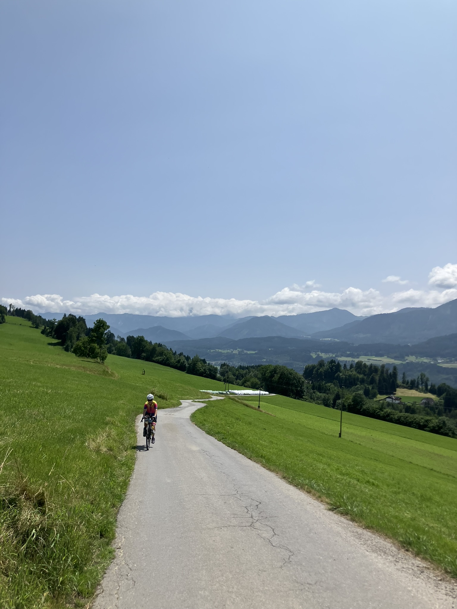 Bikepacking in Kärnten auf schmaler Asphaltstraße durch grüne Hügel mit Blick auf Alpenpanorama im Hintergrund.
