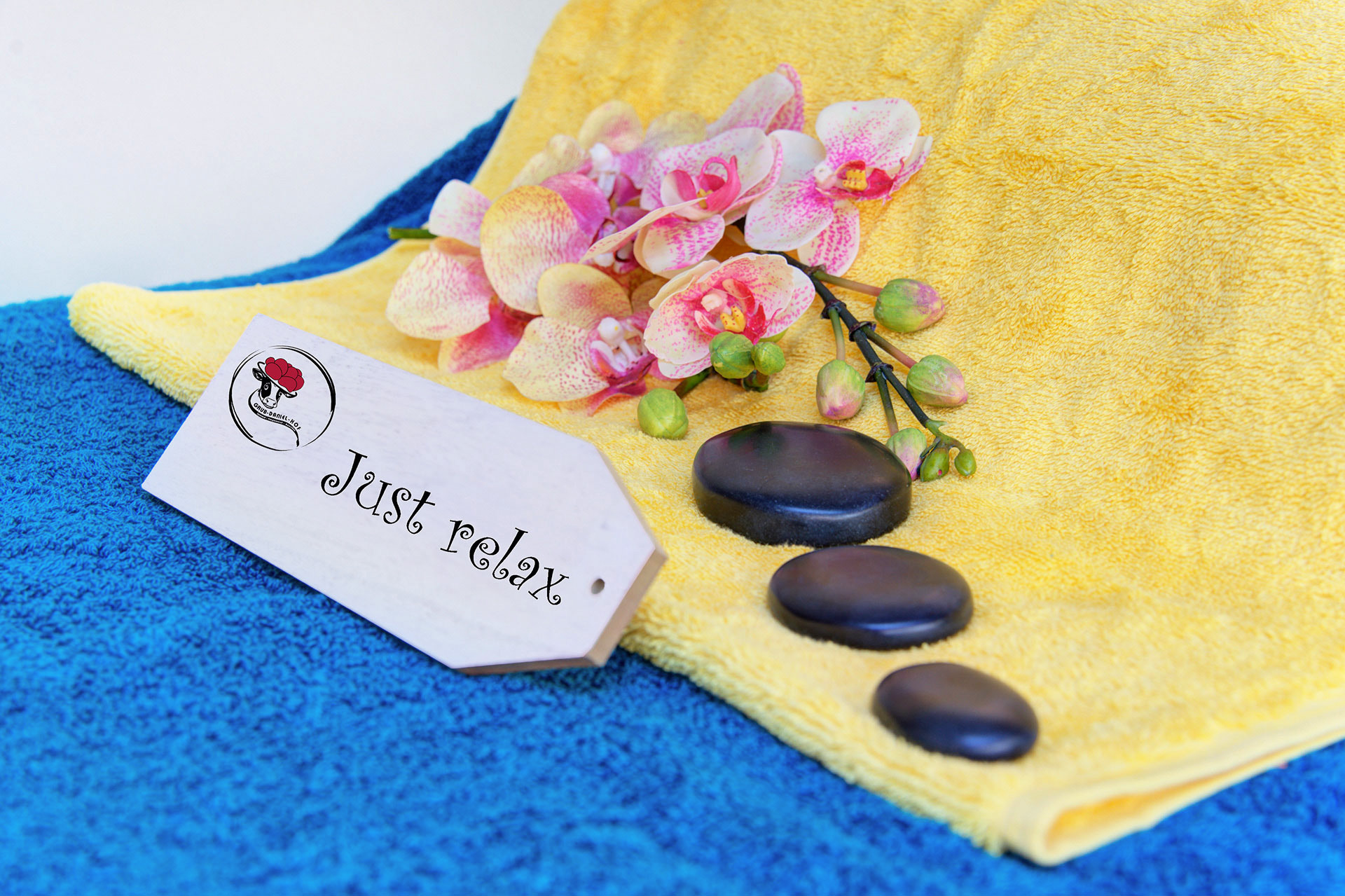 gelbes und türkises handtuch mit drei schwarzen steinen und orchideenblüte sowie schild just relax