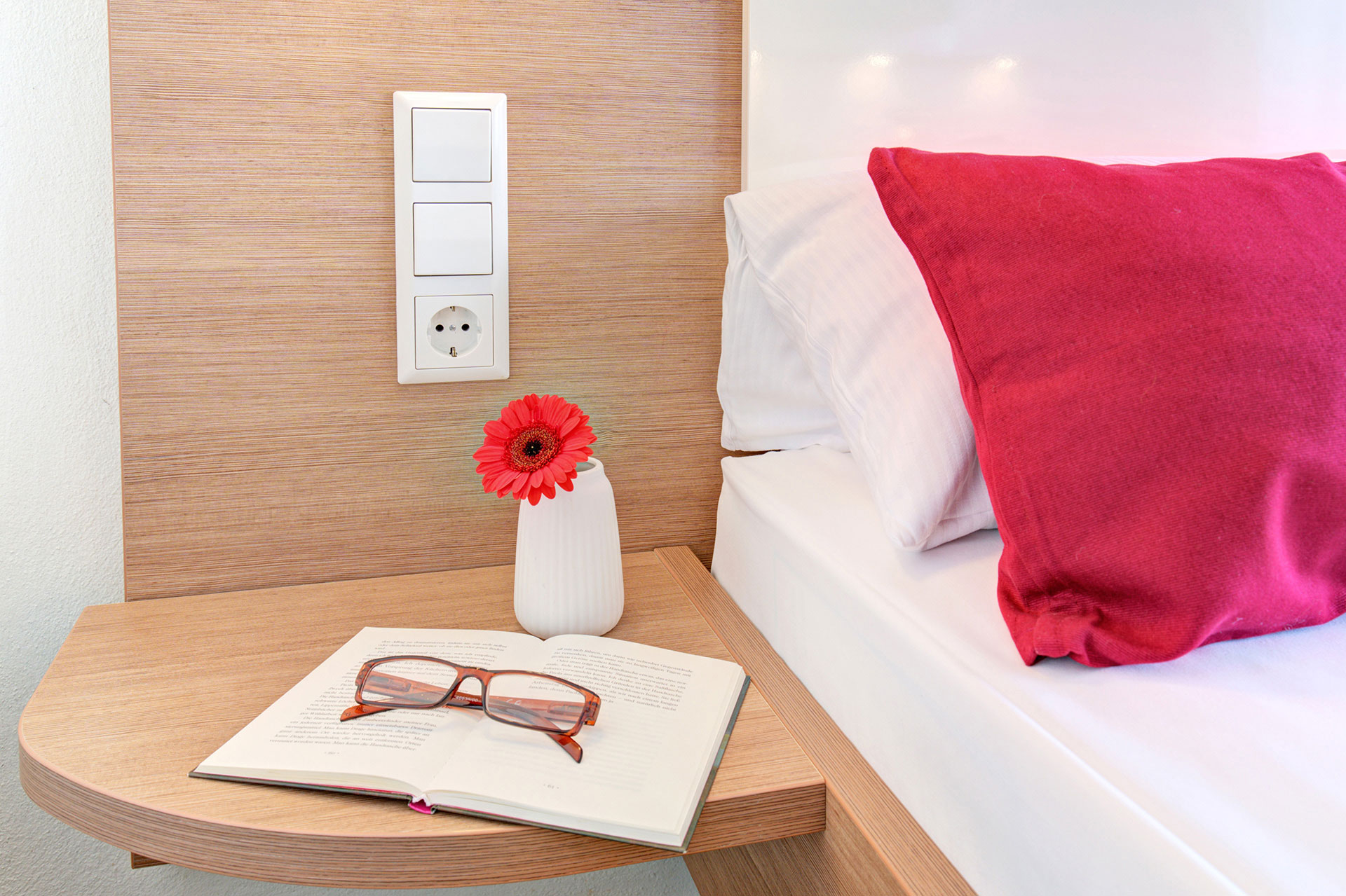nachttisch mit brille buch und rote gerbara sowie rotes kissen in hotel