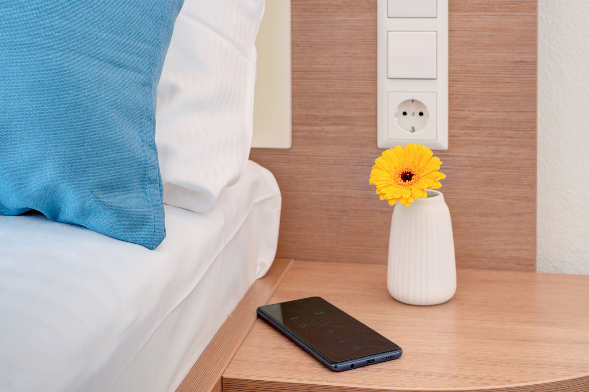 nachttisch neben türkisen kissen mit smartphone und vae mit gelber gerbara in hotel