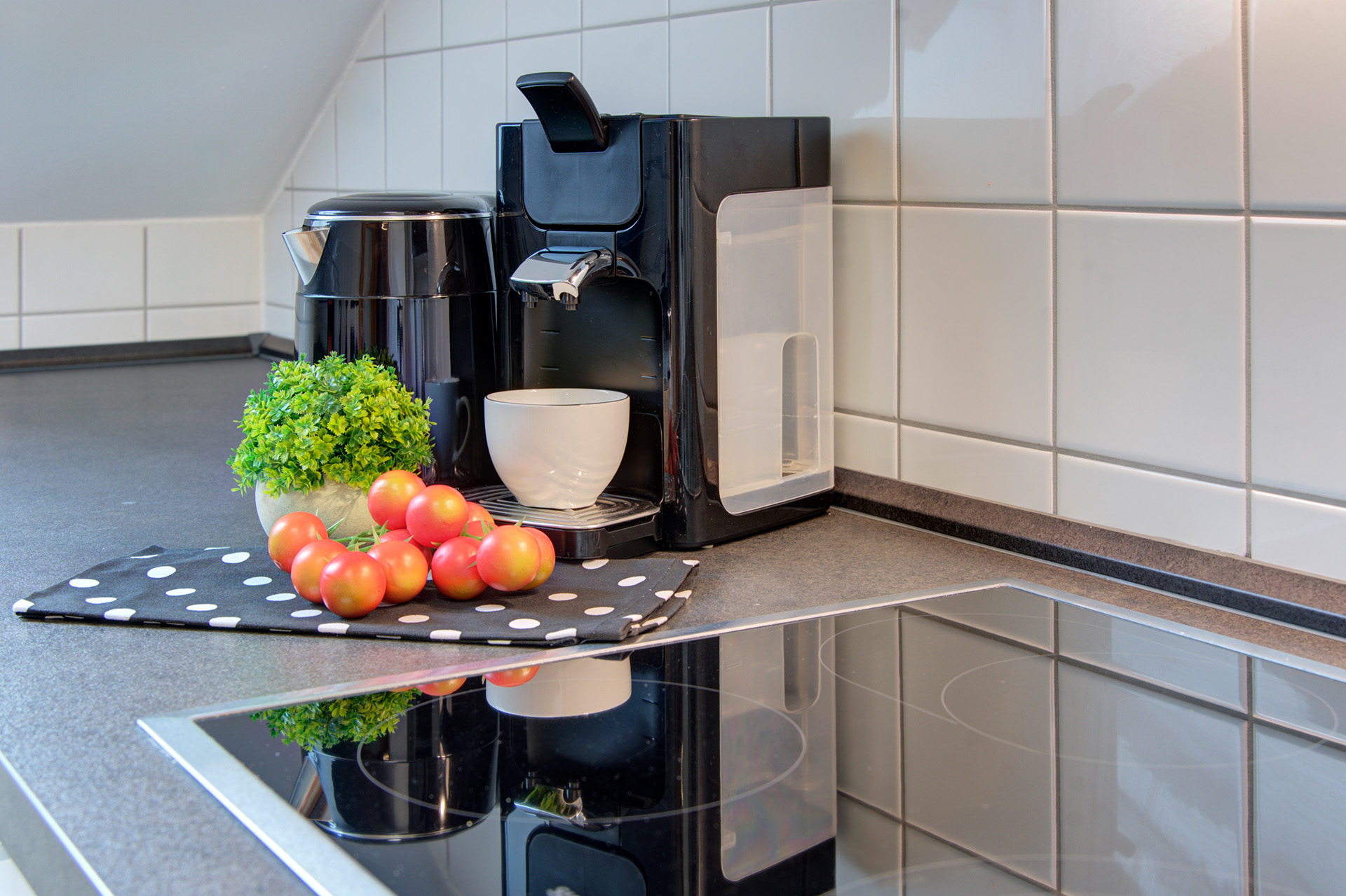 Schwarzer Wasserkocher und Espressomaschine mit Tasse sowie kleine grünpflanze und tomaten mit blick auf ceranherd