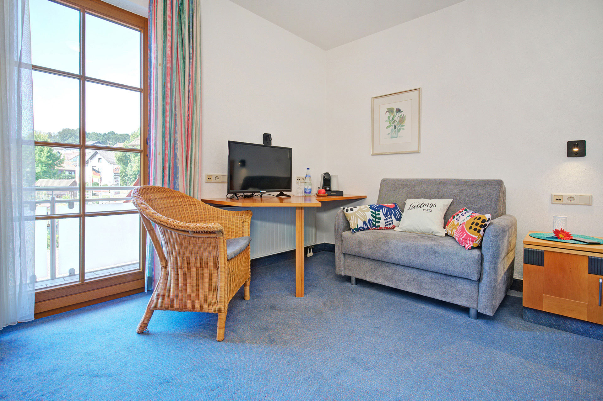 Einzelzimmer mit kleinem Sofa und TV und Korbsessel vor Balkontür