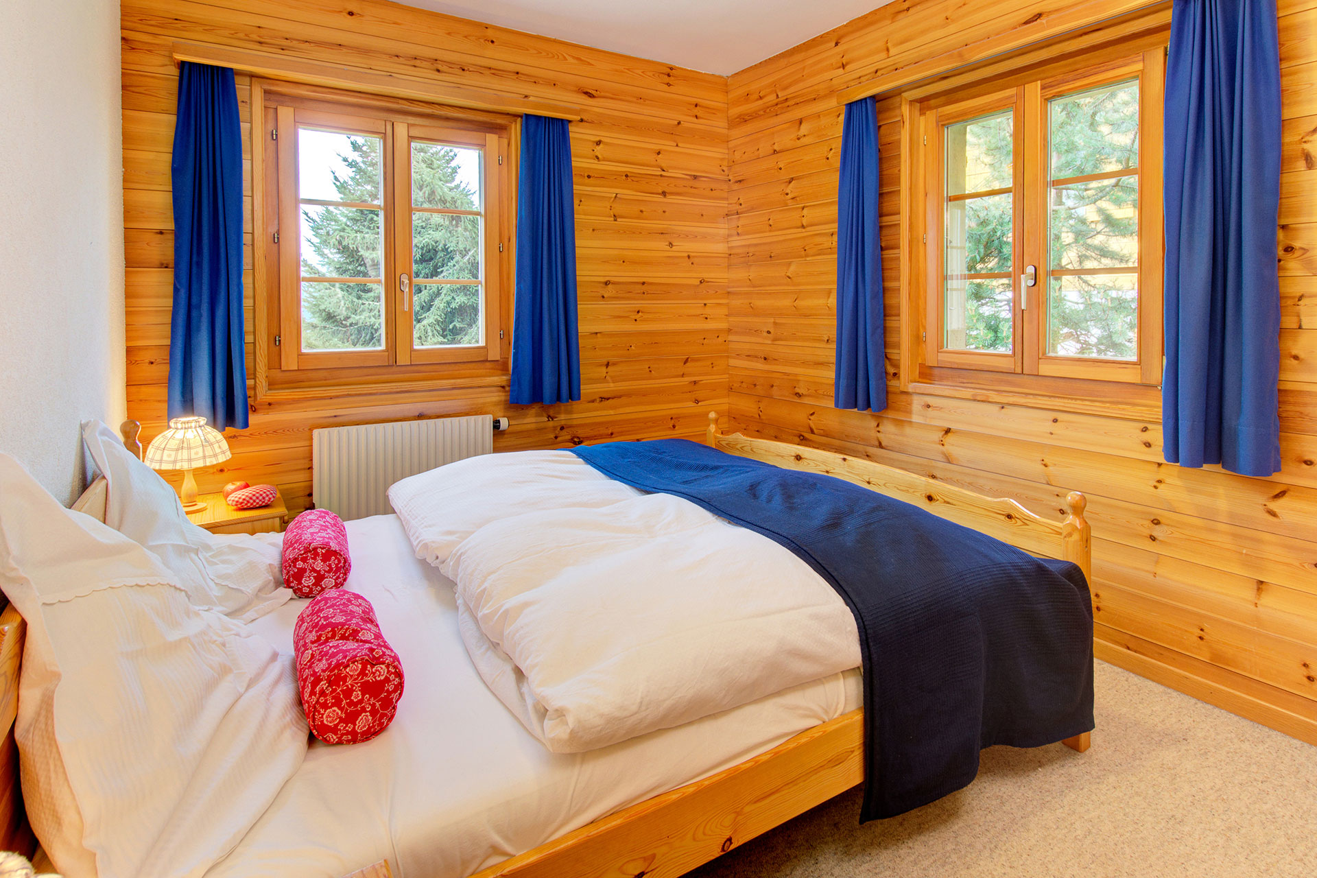 ferienhaus foto doppelzimmer mit holzwaenden mit blauem bettlaeufer und roten kleinen kissen sowie blick auf zwei fenster in den garten