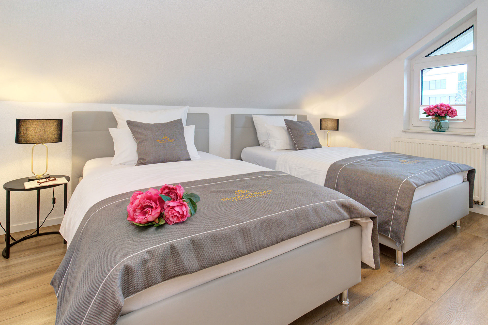 doppelzimmer mit grauen bettläufern und logo sowie lila pfingstrosen in ferienwohnung