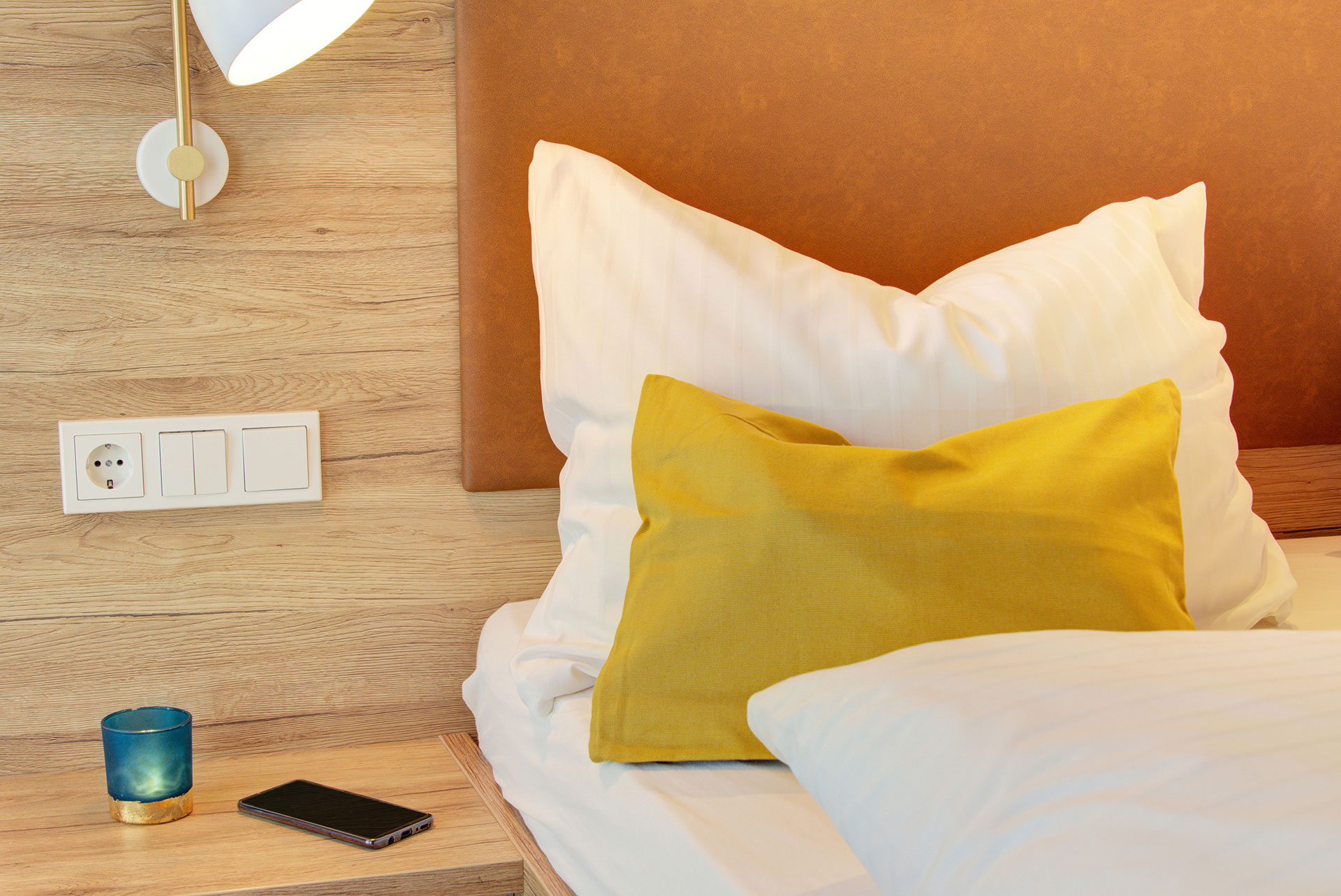 nachttisch mit smartphone und türkisem teelicht neben ockerfarbenen kissen in hotel