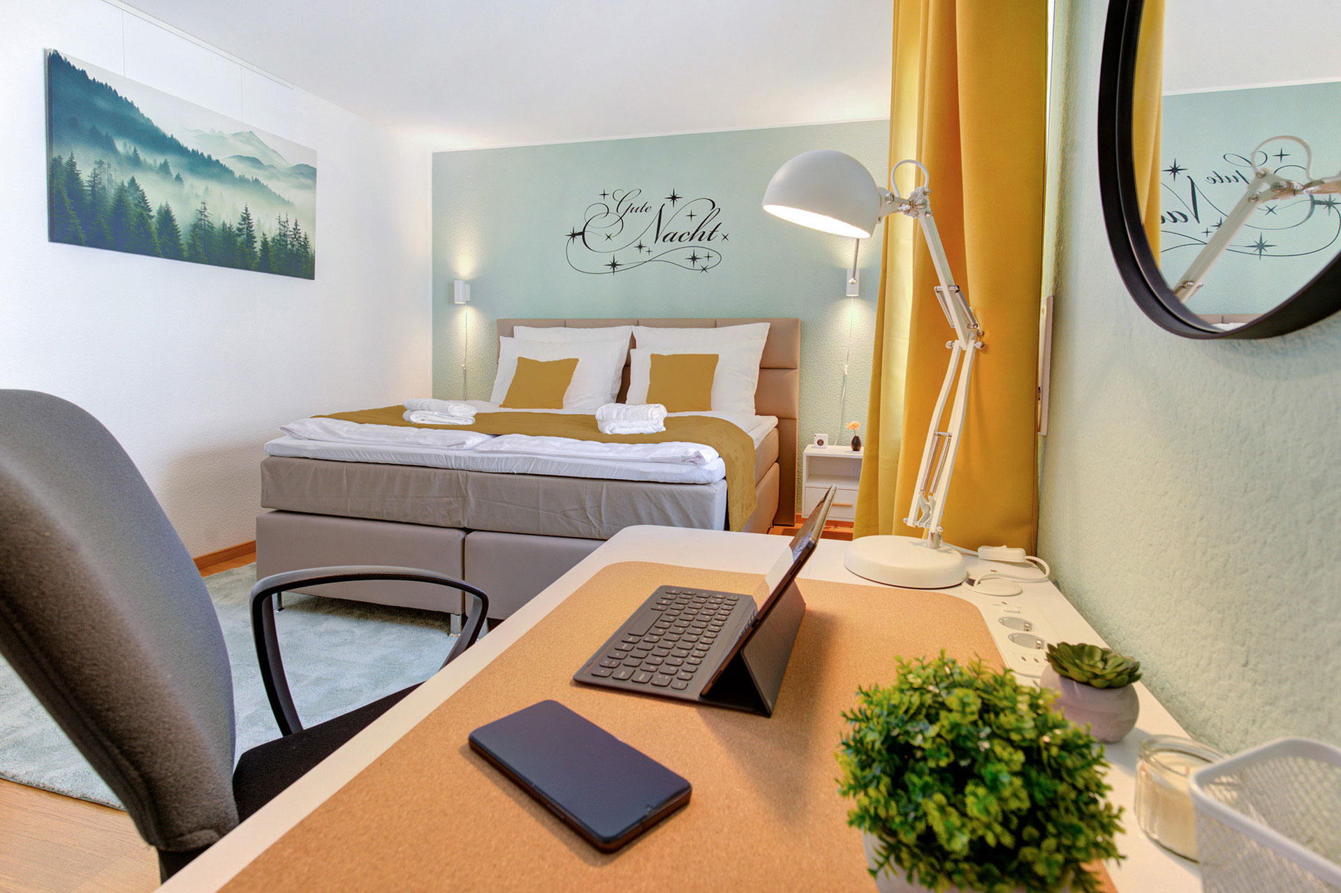 arbeitsecke in der ferienwohnung mit laptop handy und pflanze mit blick zum doppelbett über dem gute nacht steht