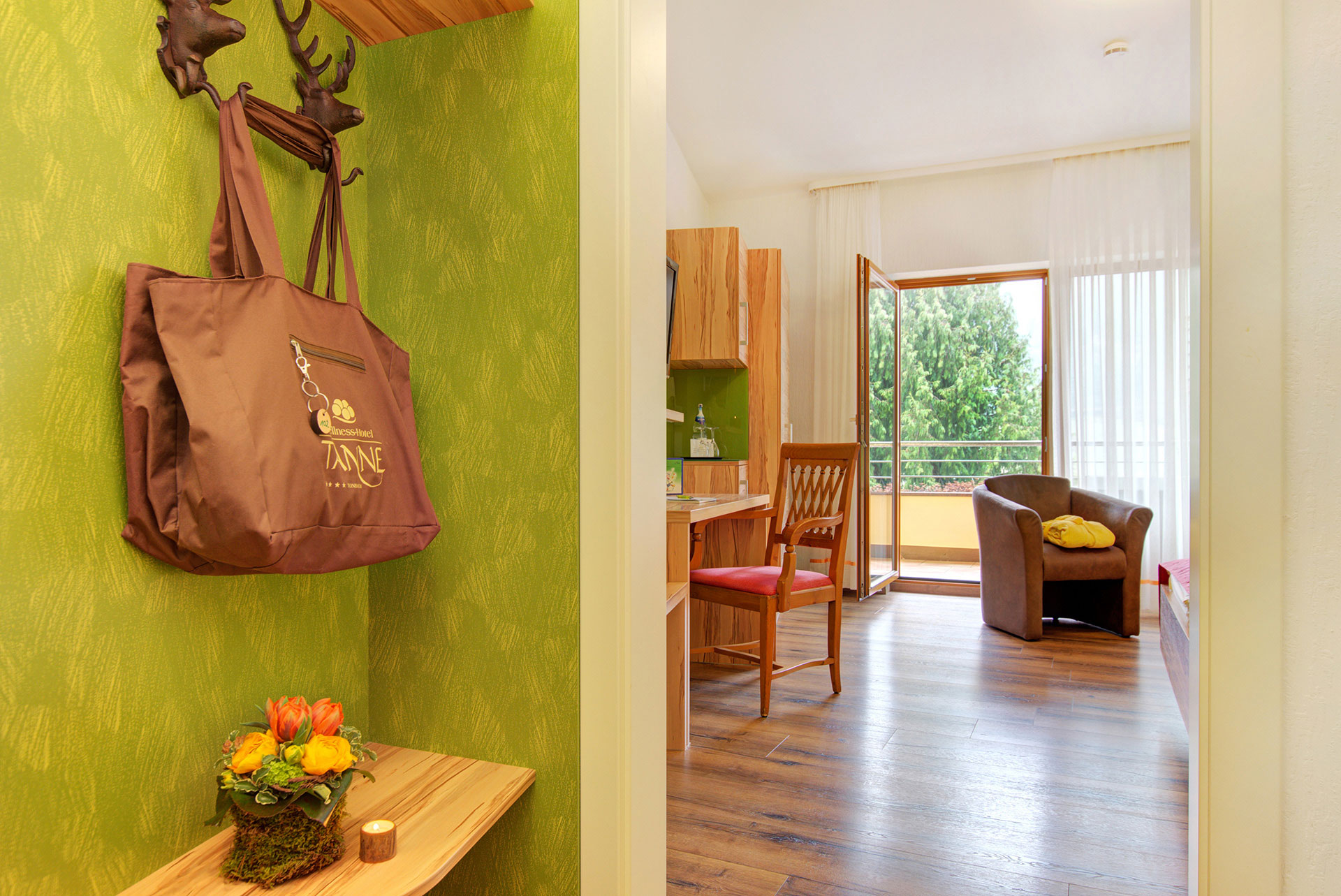 zimmereingang mit blumenbouquet und wellnesstasche sowie blick auf arbeitsbereich und balkontür in hotel