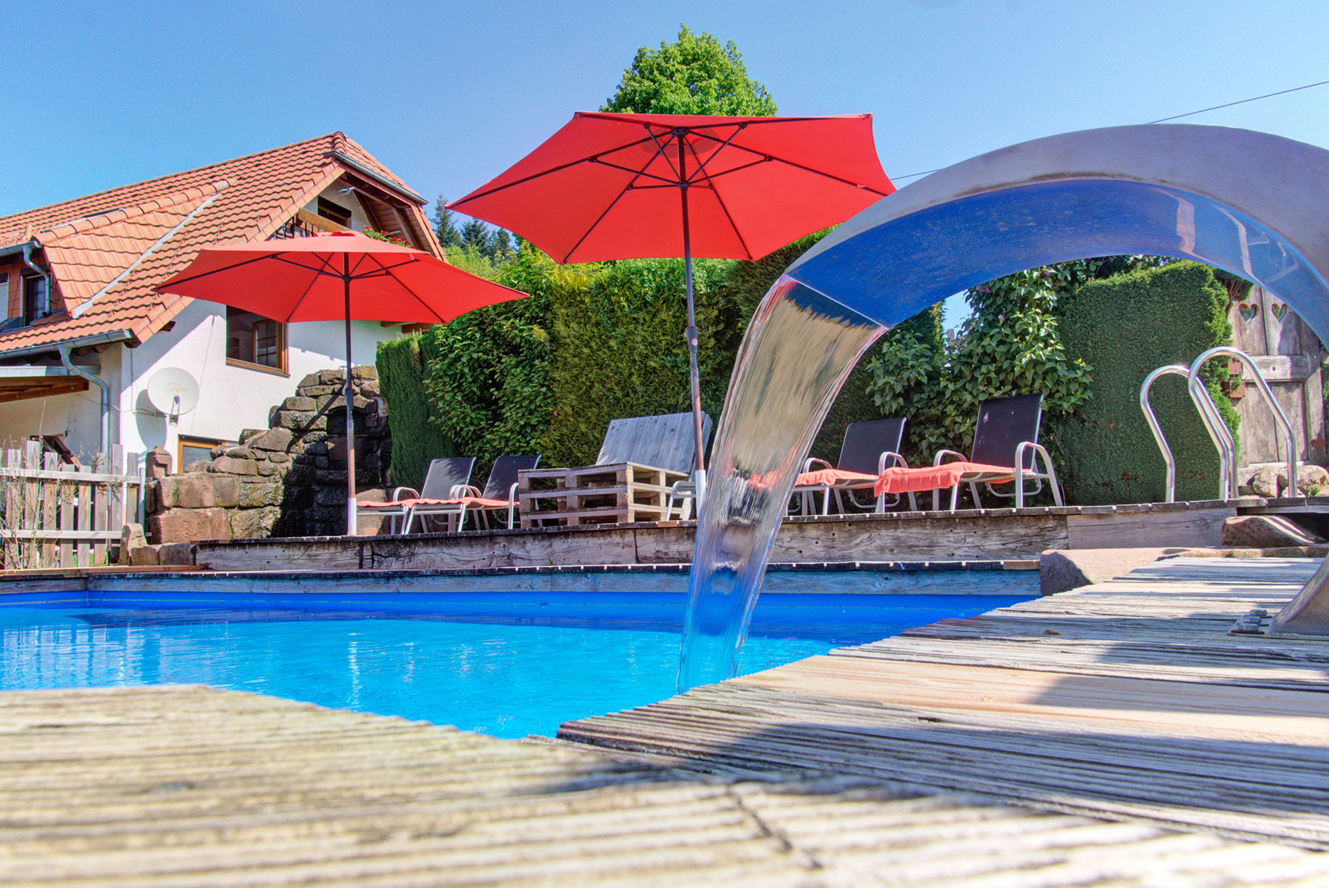 swimmingpool blick von unten auf wasserfontäne und rote schirme beim ferienhaus