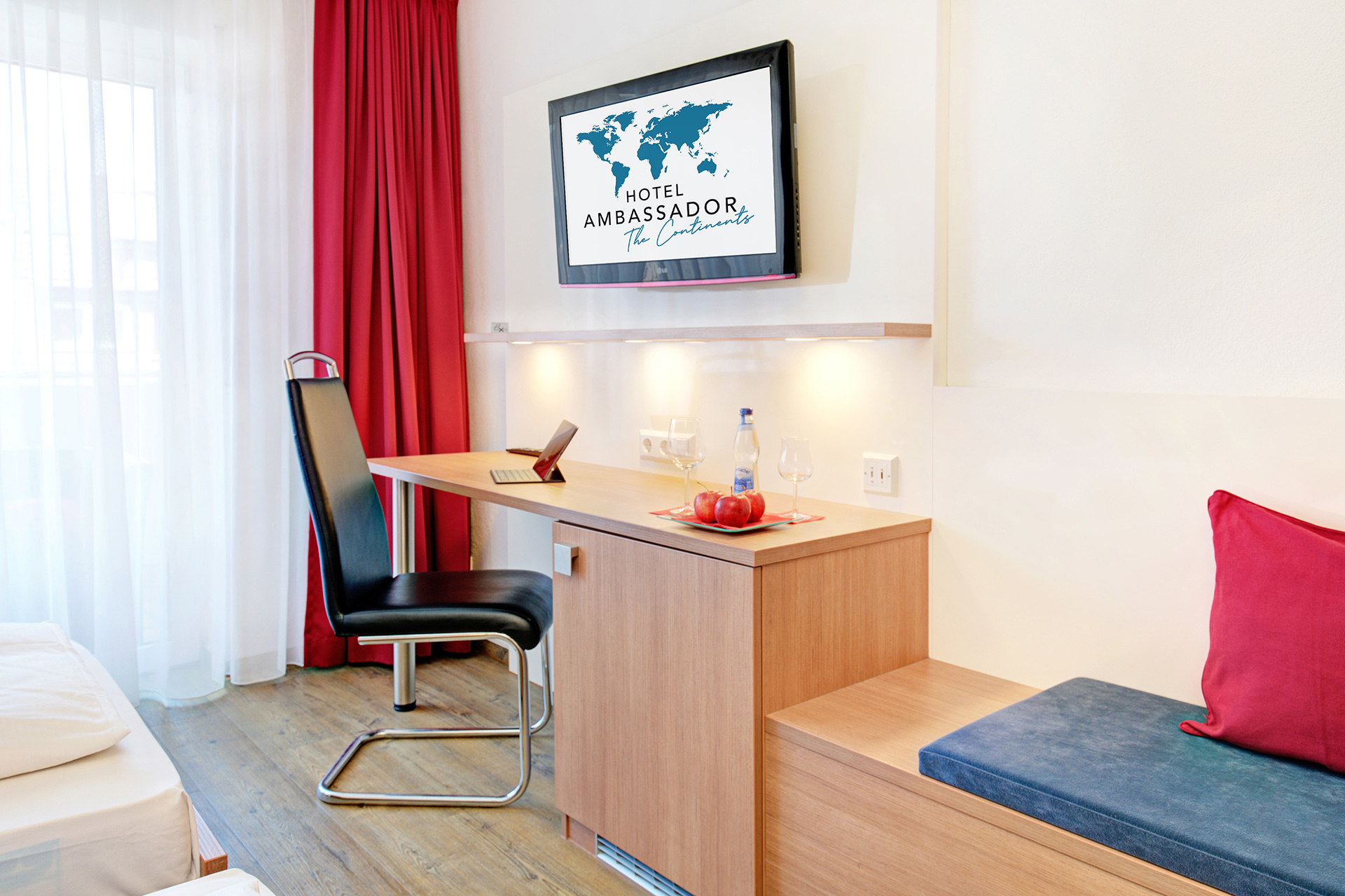 Arbeitsbereich im Hotelzimmer mit Schreibtisch und Monitor; Logo des Hotels ist auf dem Bildschirm sichtbar