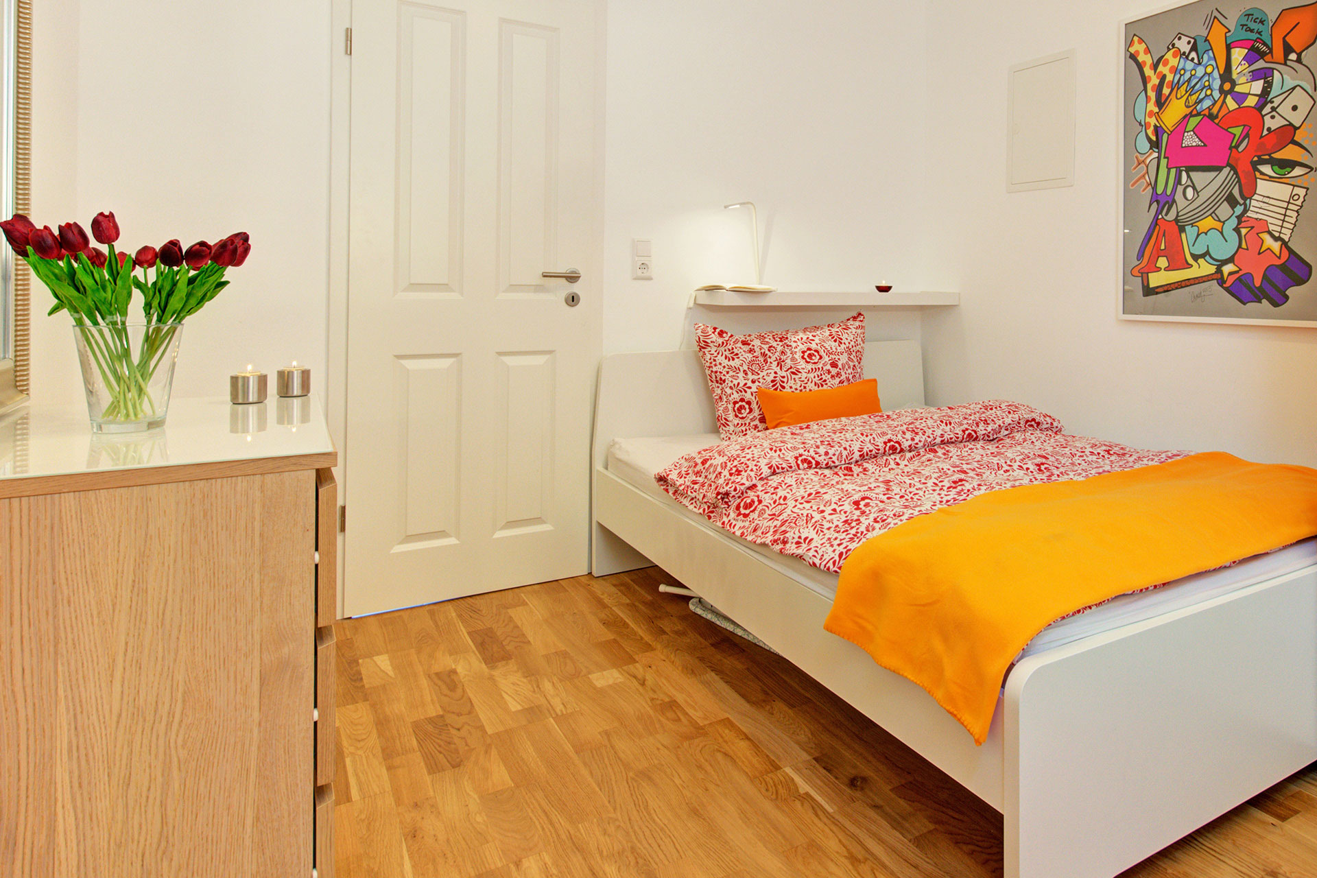 fotografin dekoriert einezlbett mit orangener decke und kissen dunkelroter tulpenstrauss auf kommode