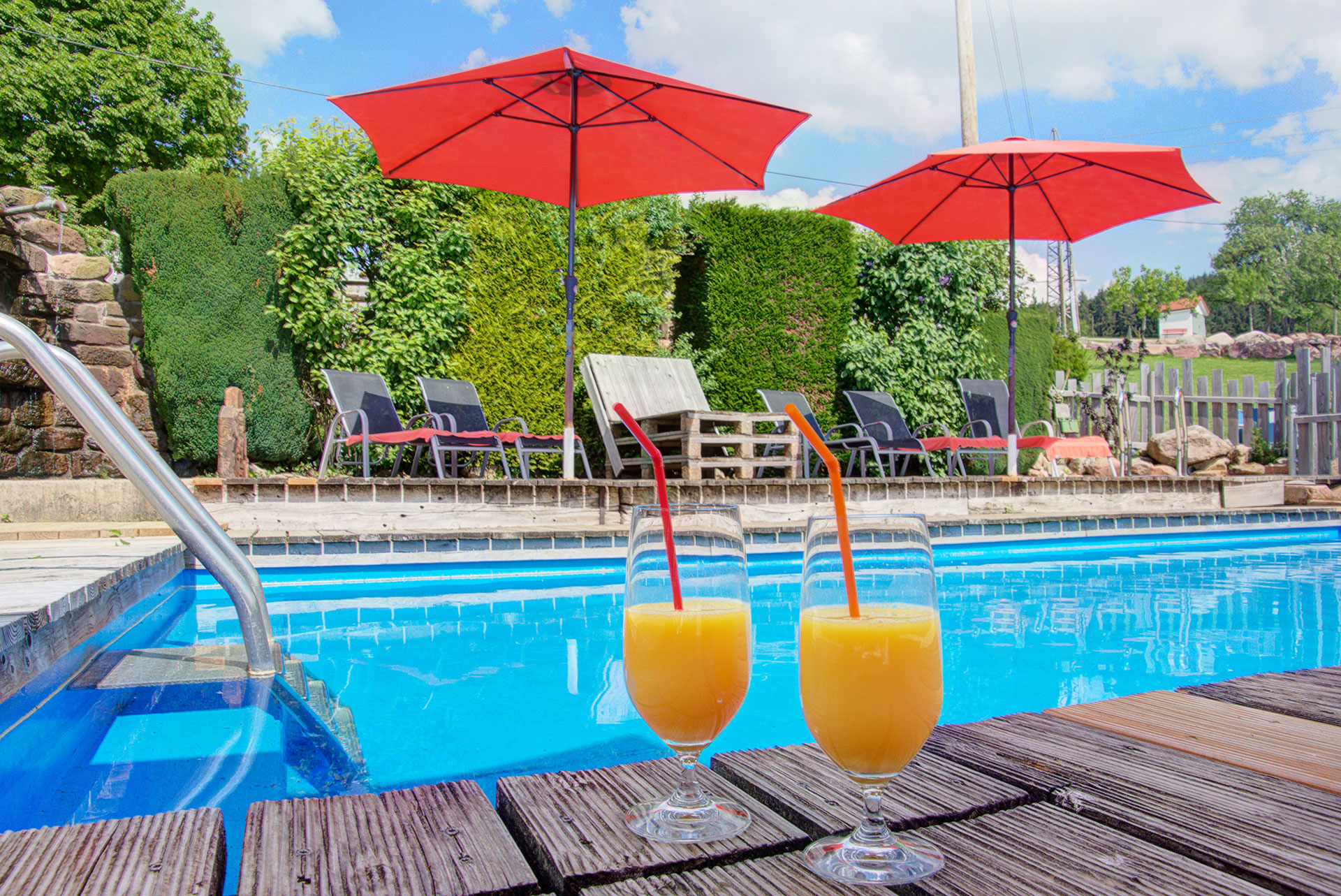 blauer swimmingpool mit cocktailorangensaftglas sowie rote schirme im hintergrund am ferienhof