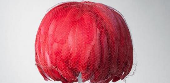 Casquete de día 1963. Casquete redondo en tafetán de rayón rojo cubierto de plumas de gallo del mismo tono. Las plumas cubren la totalidad del peinado y se encuentran protegidas por un velo de tul hexagonal en seda salmón.
