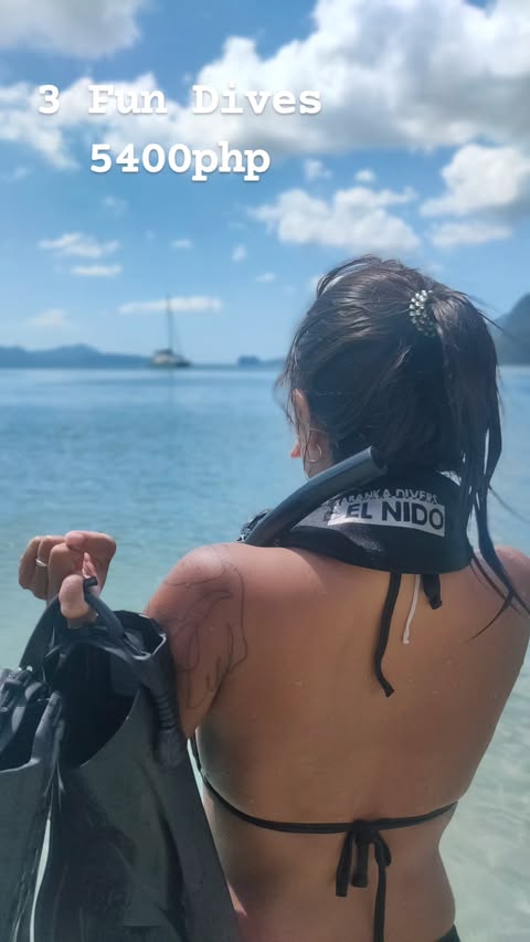 backside of a girl wearing a black bikini, holding fins, in tropical water with a sailboat in the background.