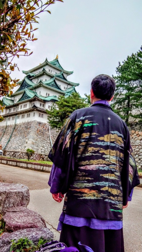 名古屋城の天守閣と石垣を背景に、手前に和モダンな羽織を着た徹平の後ろ姿が写っている写真。羽織は黒を基調に、背中には金、青、オレンジなどのカラフルで抽象的な波のような柄が描かれており、首元と裾からは紫色の着物が覗いている。左側の手前には木の葉があり、縦長の構図で構成されている。