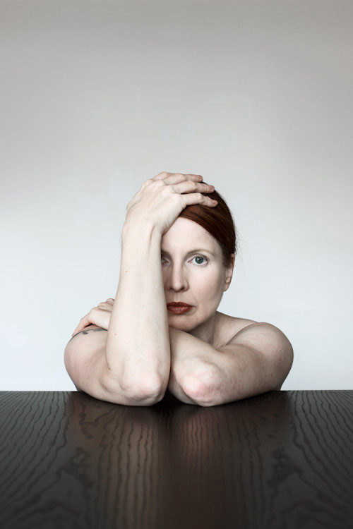 Manuela Deigert Projekte Tabletop Selbstportrait durch eine Arm halbverdecktes Gesicht in enttäuschter Haltung an einem Tisch sitzend