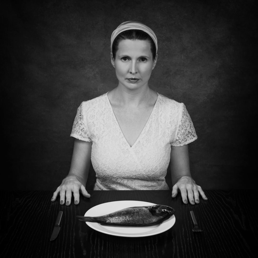 Manuela Deigert Projekte schwarzweisses Selbstportrait an einem Tisch sitzend mit einem Teller auf dem eine rohe Dorade liegt