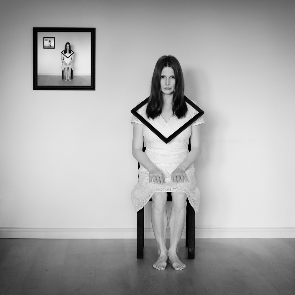 Manuela Deigert Projekte Selbstportrait mit Rahmen um den Hals auf einem Stuhl sitzend mit mehrmalig wiederholenden Selbstbild im Selbstbild an der Wand
