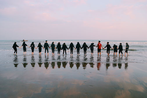 Gruppenbild am Meer, bei dem die 17 jungen Menschen durch an den Händen halten eine Kette bilden. Sie stehen barfuß im knöchelhohen Meer.ser