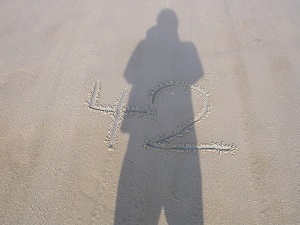 Diese Farbfotografie von Thorsten Hülsberg zeigt seinen Schatten an einem dänischen Sandstrand, wo eine 42 geschrieben steht.