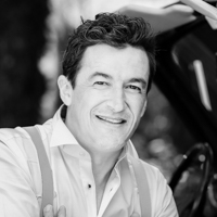 Homme souriant en noir et blanc, portant une chemise blanche et des bretelles, avec un fond flou en extérieur.