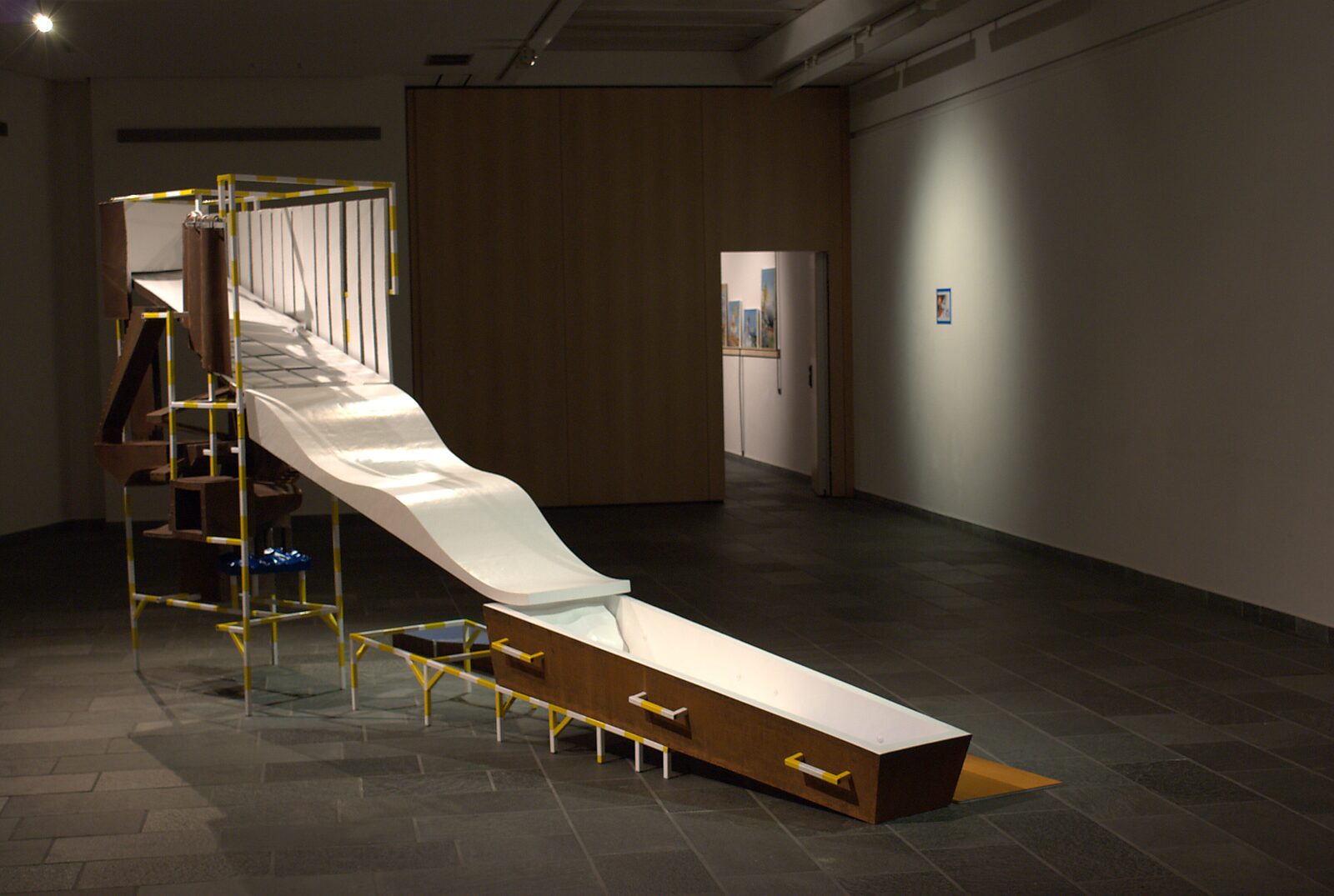 ‘Pro-Life’: Tentoonstellingszicht met lange witte glijvorm die uitmondt in een open kist; metalen structuur en houten panelen op de achtergrond, CC De Adelberg, Lommel, 2015.